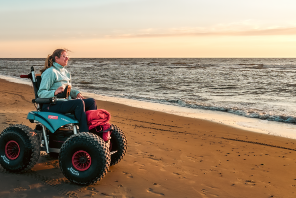 April Ranshuijsen zitten in elektrische voortgedreven strandrolstoel met grote off the road wielen. Zij staat aan de waterlijn en kijkt uit over de zee.