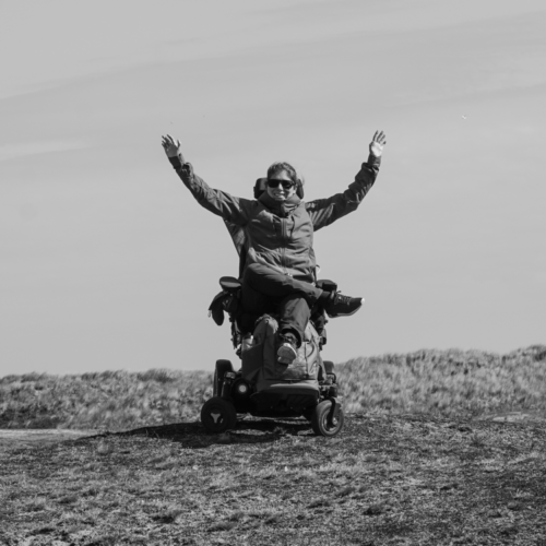 Foto van April in haar rolstoel op een open veld. Ze heeft haar handen in de lucht en kijkt naar de camera.