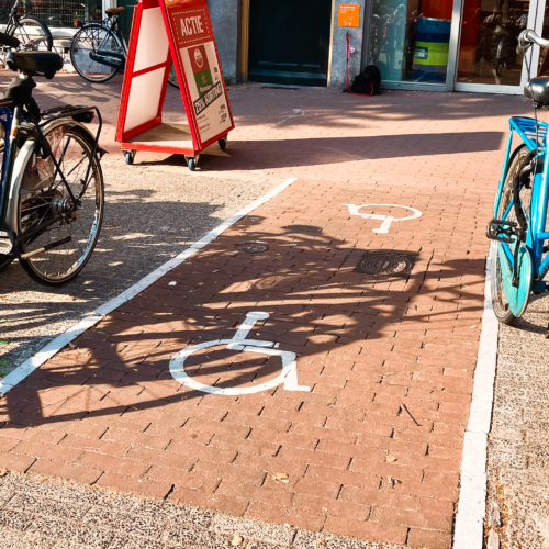 Tussen twee fietsenrekken met fietsen in lopen twee wit geverfde lijnen die ene pad vormen. Op de rode stenen van het pad staat het symbool van een rolstoel geverfd.