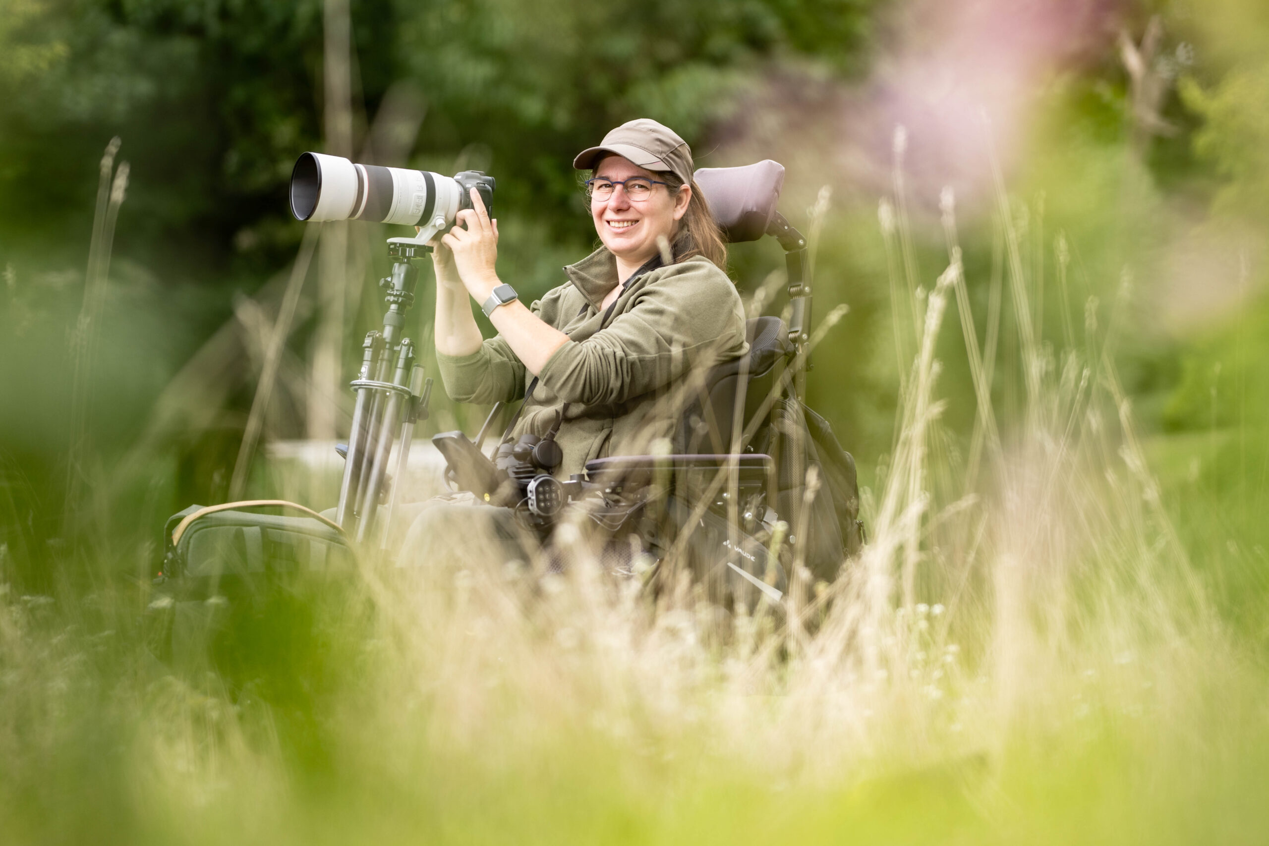 April Ranshuijsen zit in haar rolstoel met voorop een camera met telelens. Zij kijkt zijwaarts naar de fotograaf tussen dromerige grashalmen door.