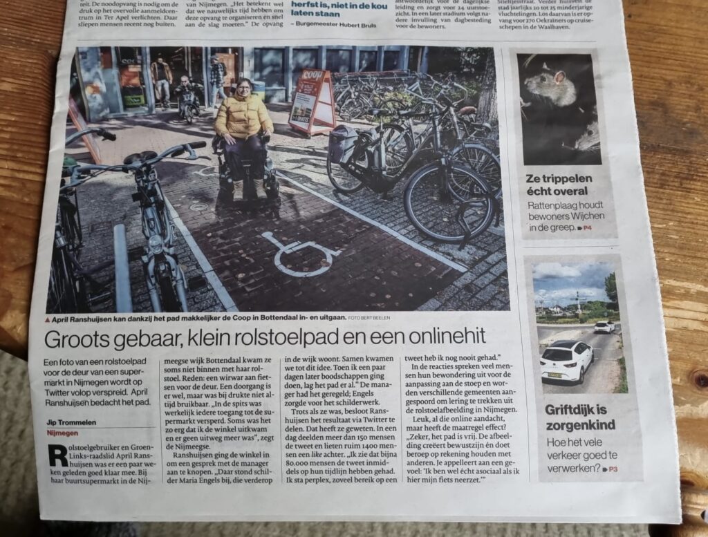 Foto van de voorpagina van De Gelderlander. Daarop staat April in haar rolstoel op een met lijnen afgebakend pad tussen twee fietsenrekken door. Op de stenen van de straat is in wit het rolstoel symbool groot geschilderd.